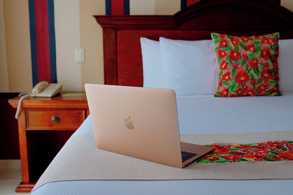 Una laptop sobre una cama con una colcha estampada, una mesita de noche de madera con un teléfono, y una pared a rayas azules y rojas.