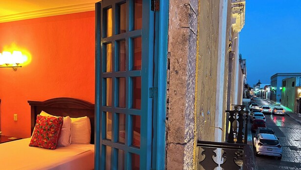 A hotel room with a bed, a blue door, and a view of a street with cars. - Hotel Plaza Colonial (Campeche)