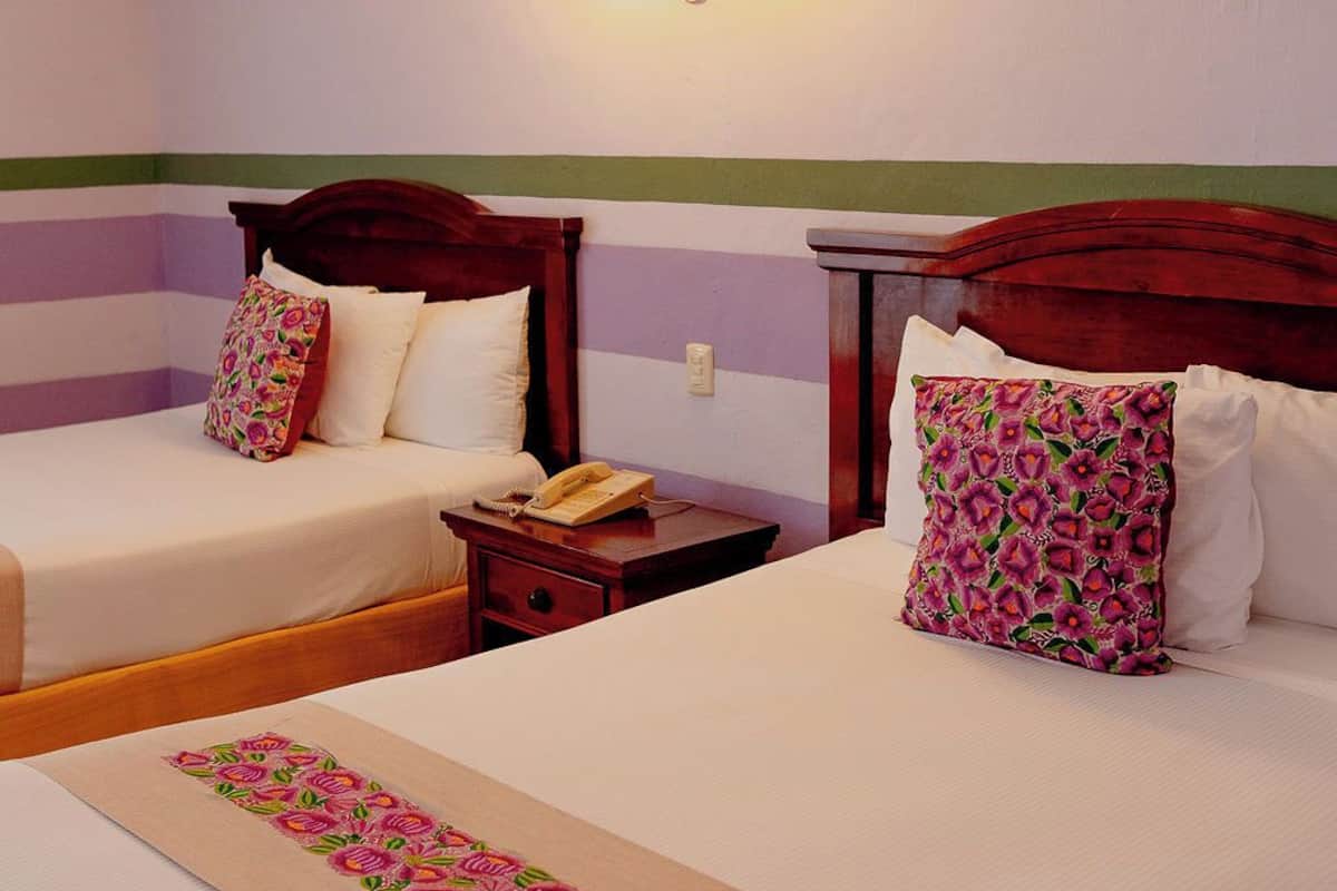 Dos camas con almohadas estampadas con motivos florales, una mesita de noche con teléfono, y un aire acondicionado instalado en la pared.