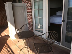 A patio with two metal chairs and a small table, a sliding glass door leading to a bedroom with a bed and a nightstand. - The Glen Club (Glenview)