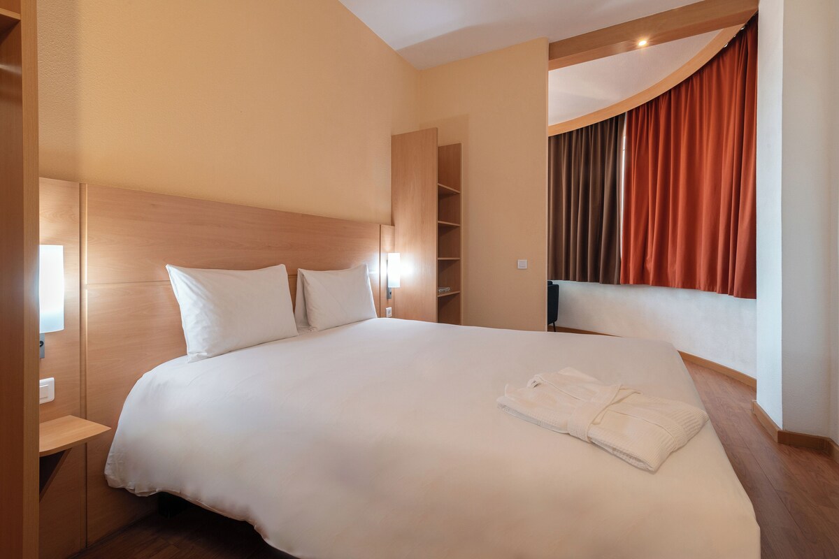 A hotel room with a large bed, white bedding, a wooden headboard, a built-in shelf, and a window with curtains.
