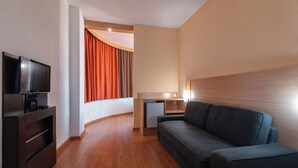 Una habitación de hotel moderna con un sofá de color oscuro, un televisor de pantalla plana, un escritorio pequeño con una lámpara y un refrigerador compacto.