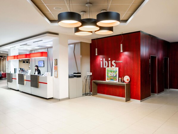 Un mostrador de recepción con una pared roja y un letrero enmarcado que dice 'ibis', un techo moderno con luces colgantes y un piso de baldosas limpias.