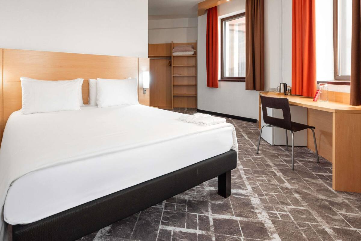 Une chambre d’hôtel avec un grand lit, un bureau, une chaise, une armoire et une fenêtre avec des rideaux.