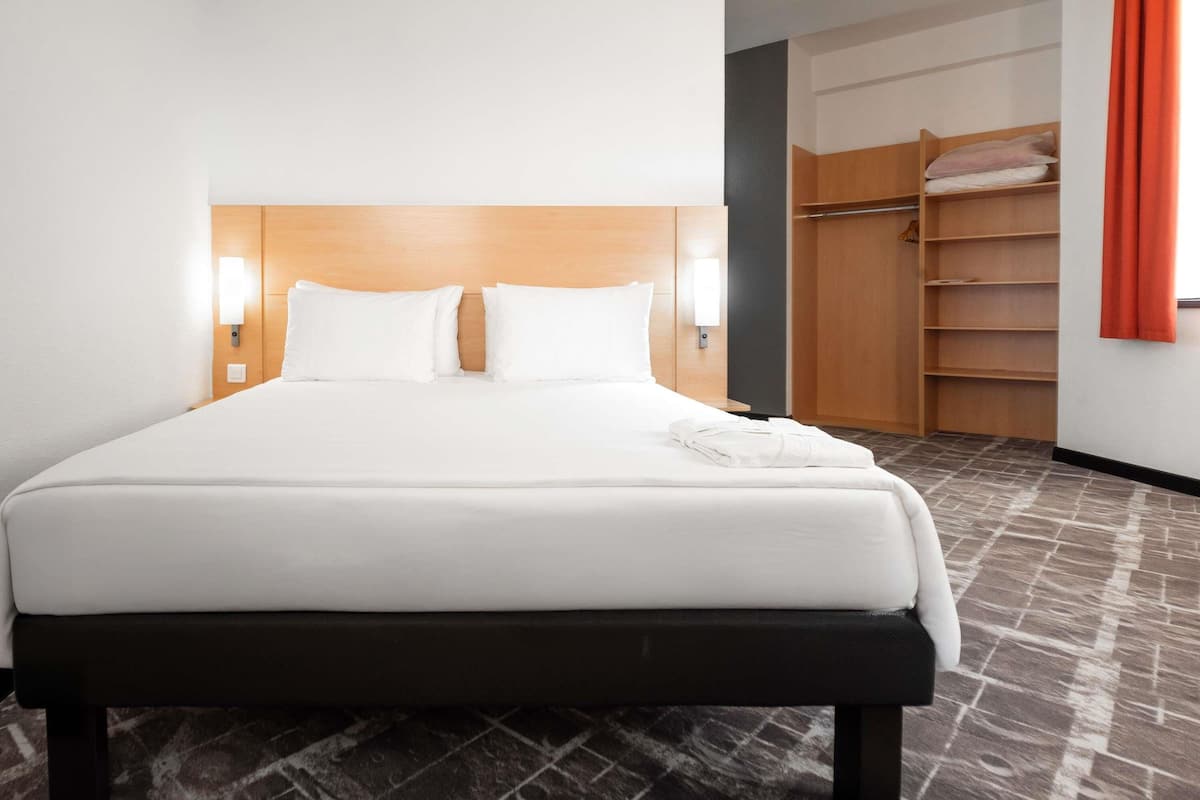 Une chambre d’hôtel avec un lit, des lampes de chevet, une tête de lit en bois, une armoire avec des étagères et une moquette à motifs.