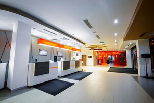 Zona de recepción moderna de un hotel, con un mostrador de recepción, una pantalla digital y un restaurante con decoración roja al fondo.