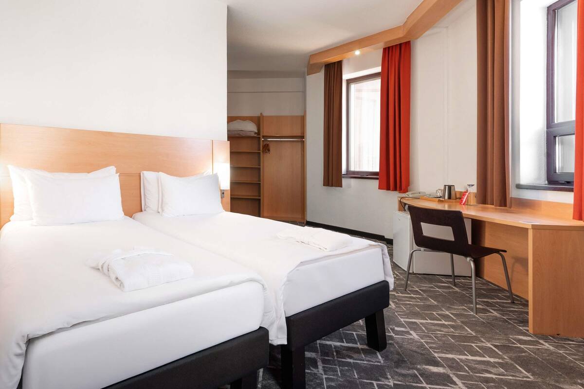 Habitación de hotel con dos camas, un escritorio, una silla, un armario y un ventanal con cortinas.