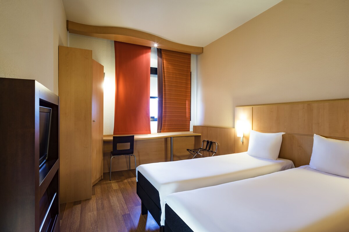 A hotel room with two beds, a desk, a chair, and a TV.