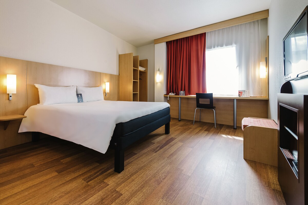 A hotel room with a bed, desk, chair, wardrobe, and a TV.