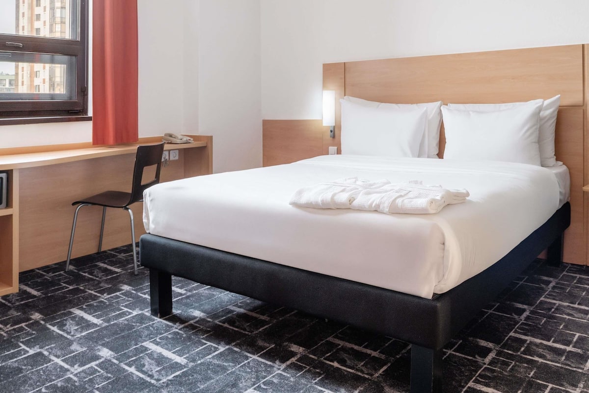 Une chambre d’hôtel équipée d’un lit, d’un bureau, d’une chaise et d’une fenêtre donnant sur des immeubles.
