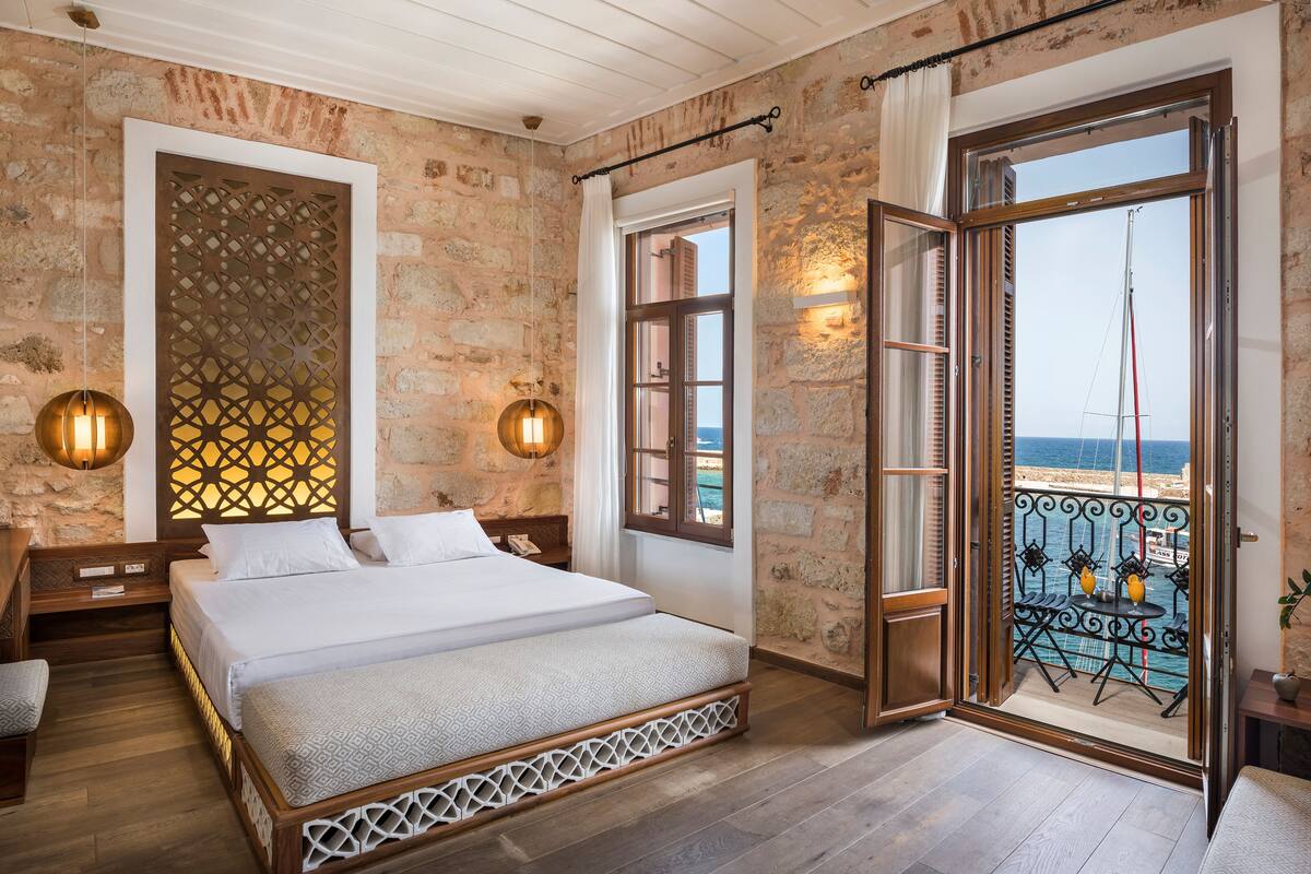 A bedroom with a large bed, a wooden headboard, a nightstand, and a view of the sea through an open door.