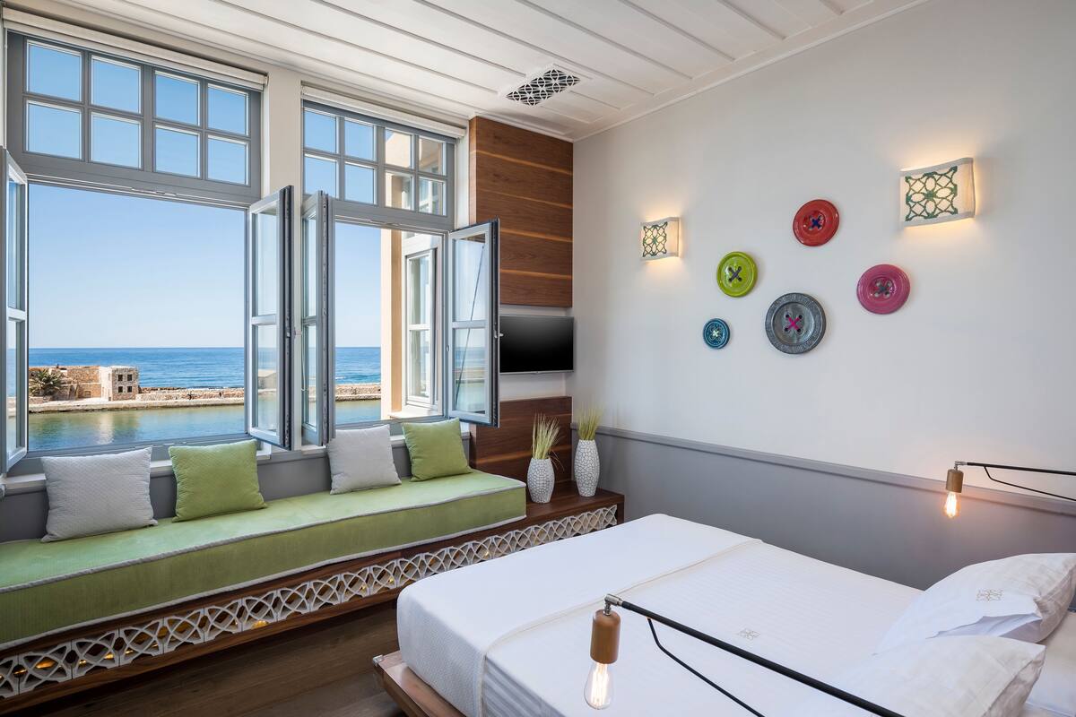 A bedroom with a large window, a bed, a seating area with cushions, and decorative wall plates.