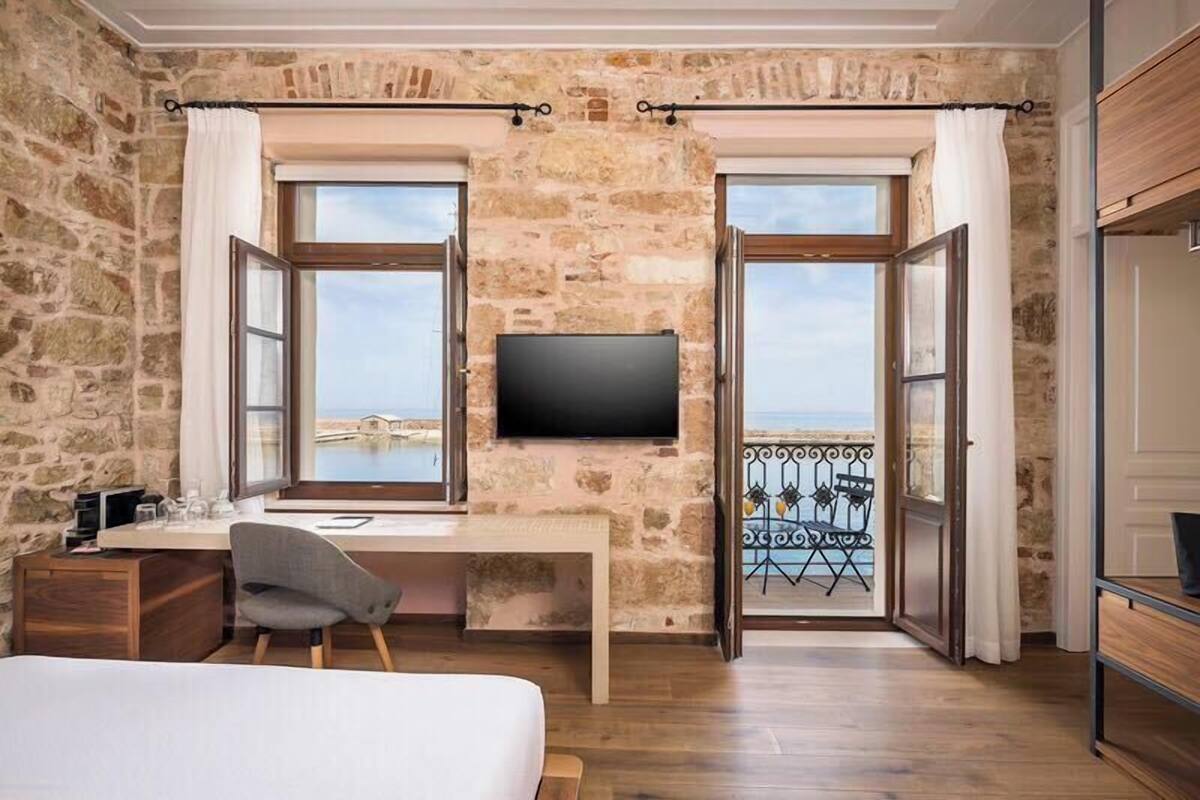 A room with a stone wall, a desk with a chair, a bed, and a TV mounted on the wall.