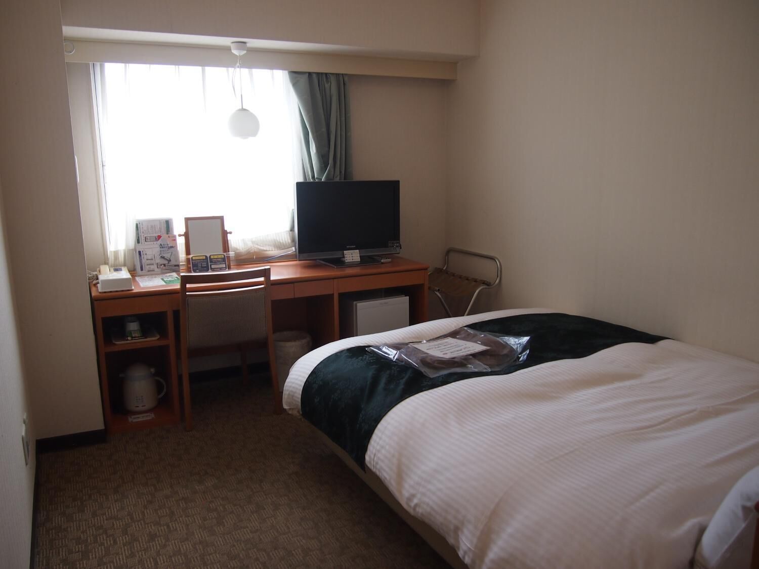 A hotel room with a bed, a desk with a computer, a chair, a small table with a coffee maker, and a window with curtains.