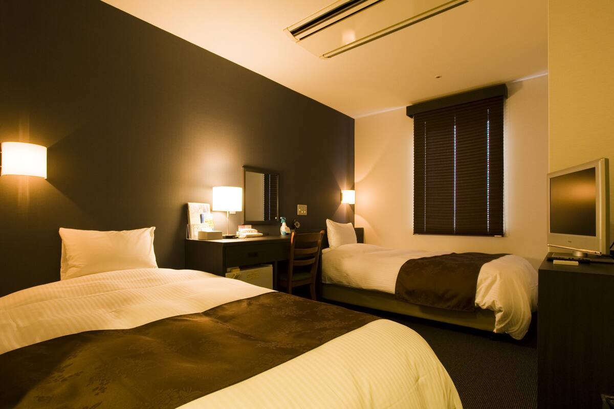 Une chambre d’hôtel avec deux lits, un bureau, une chaise, une télévision et une fenêtre avec des stores.