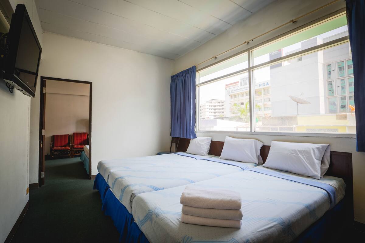 A hotel room with two beds, a TV, a window with curtains, and a view of buildings outside.