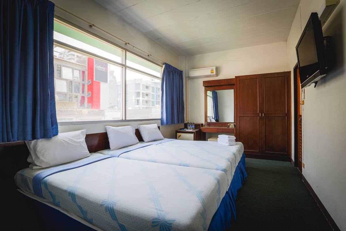 A hotel room with two beds, a large window with curtains, a wooden wardrobe, a flat-screen TV, and a bathroom with a mirror and sink.