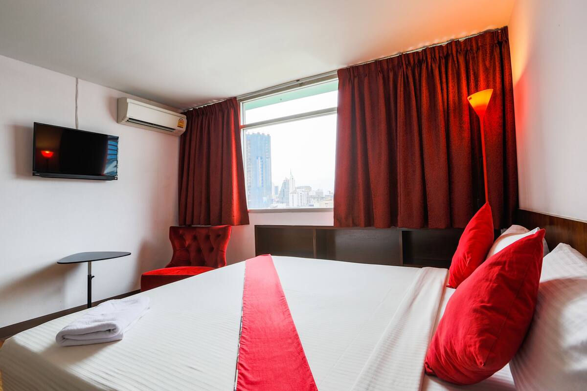 A hotel room with a bed, a TV mounted on the wall, a window with curtains, and a city view.
