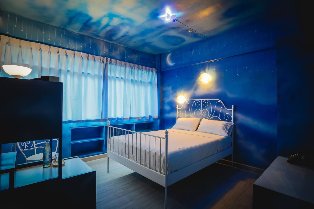 A bedroom with a bed, a bedside table, a lamp, and a night sky mural on the ceiling.