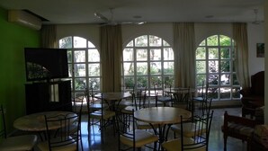 Un comedor con varias mesas y sillas, amplias ventanas en arco y un televisor sobre un mueble.