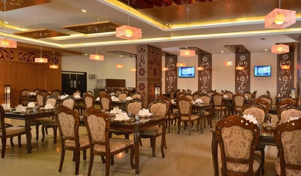 A large banquet hall with multiple dining tables and chairs, ornate wall decorations, and ceiling lights.