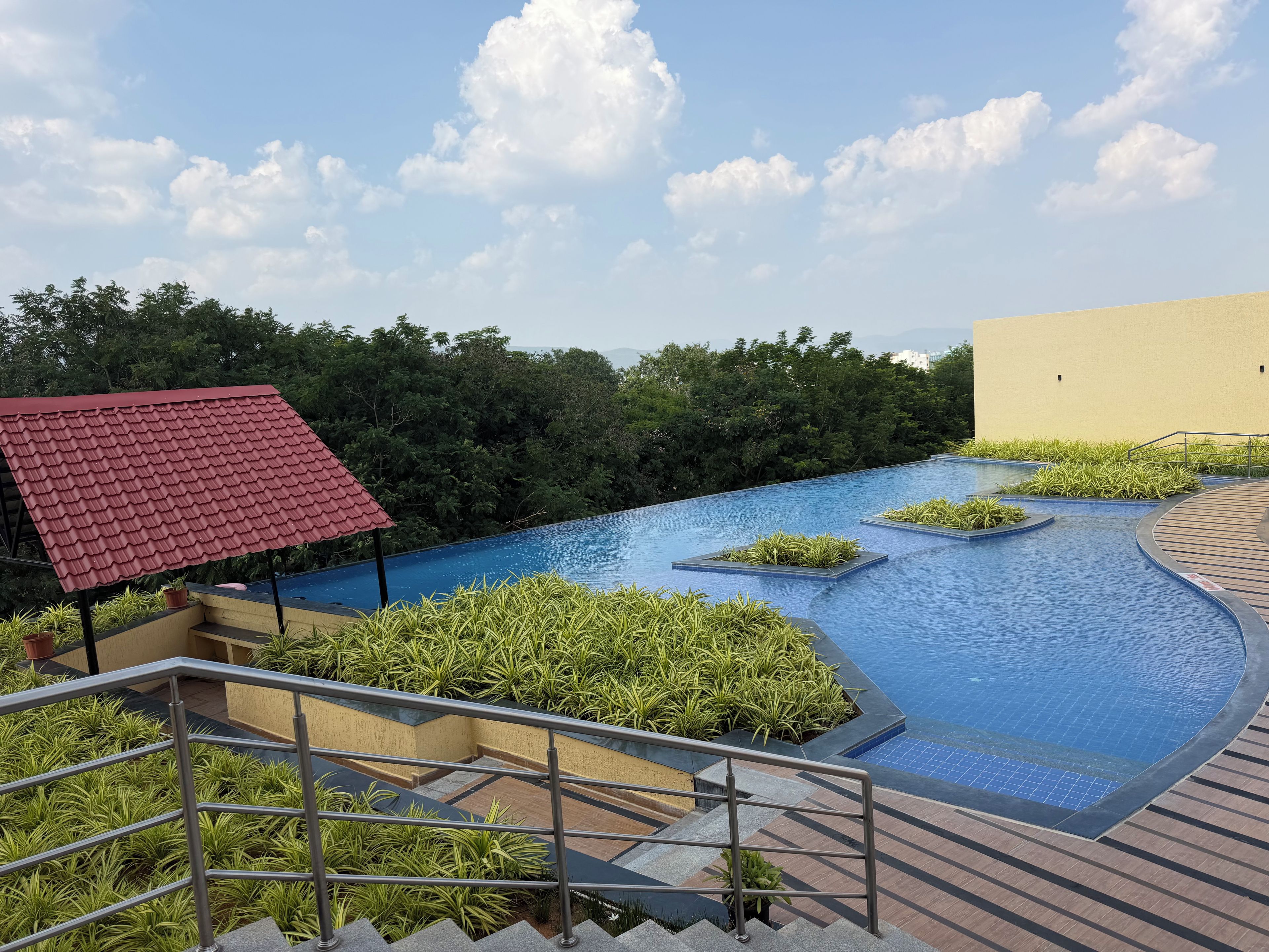 A rooftop infinity pool with a red-roofed structure, surrounded by lush greenery and a clear sky.