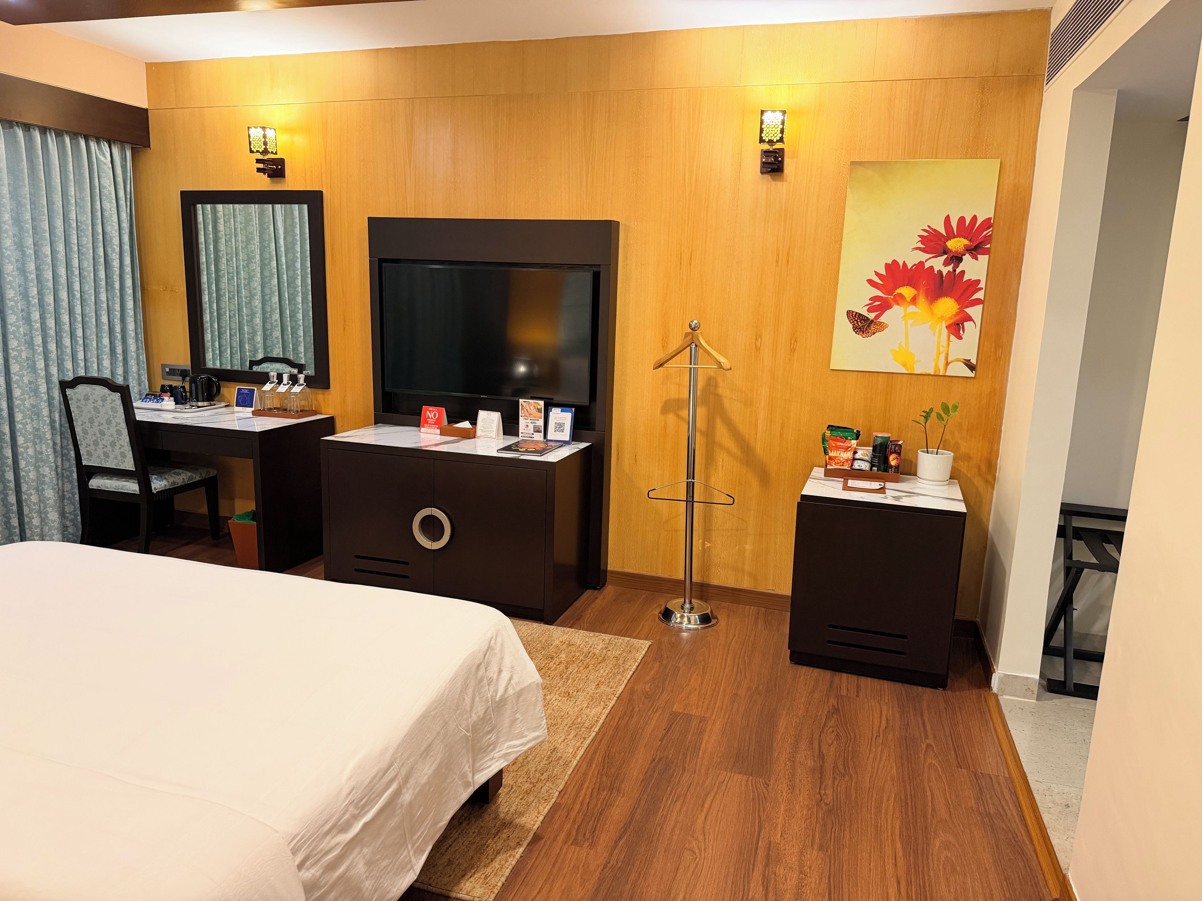 Habitación de hotel con cama, escritorio, televisor, espejo, lámpara de pared y un cuadro.