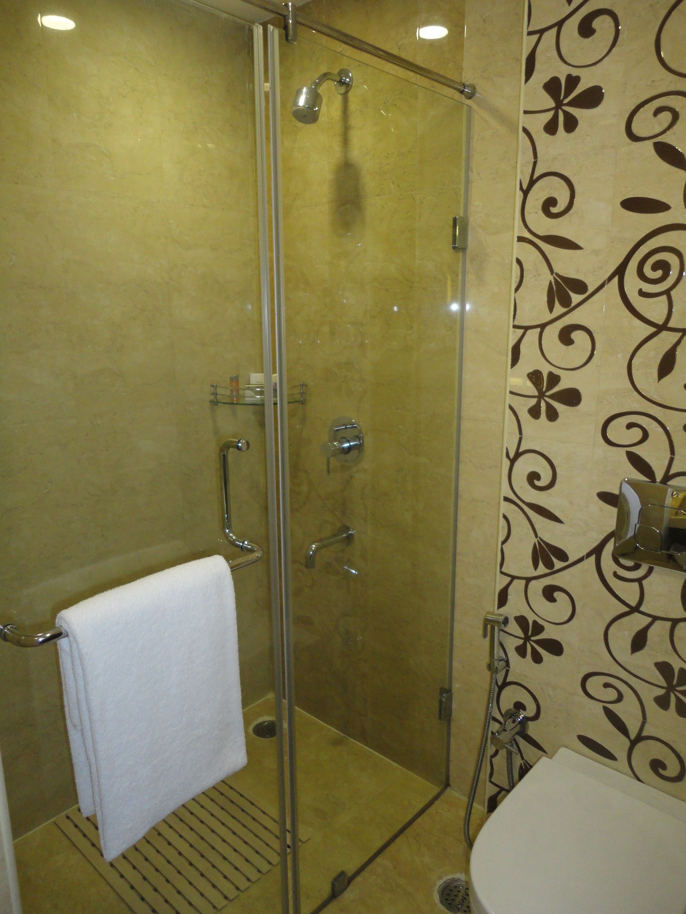 Ducha con cerramiento de vidrio, cabezal de ducha de mano, una toalla blanca colgada en un perchero metálico y un lavabo con grifo.