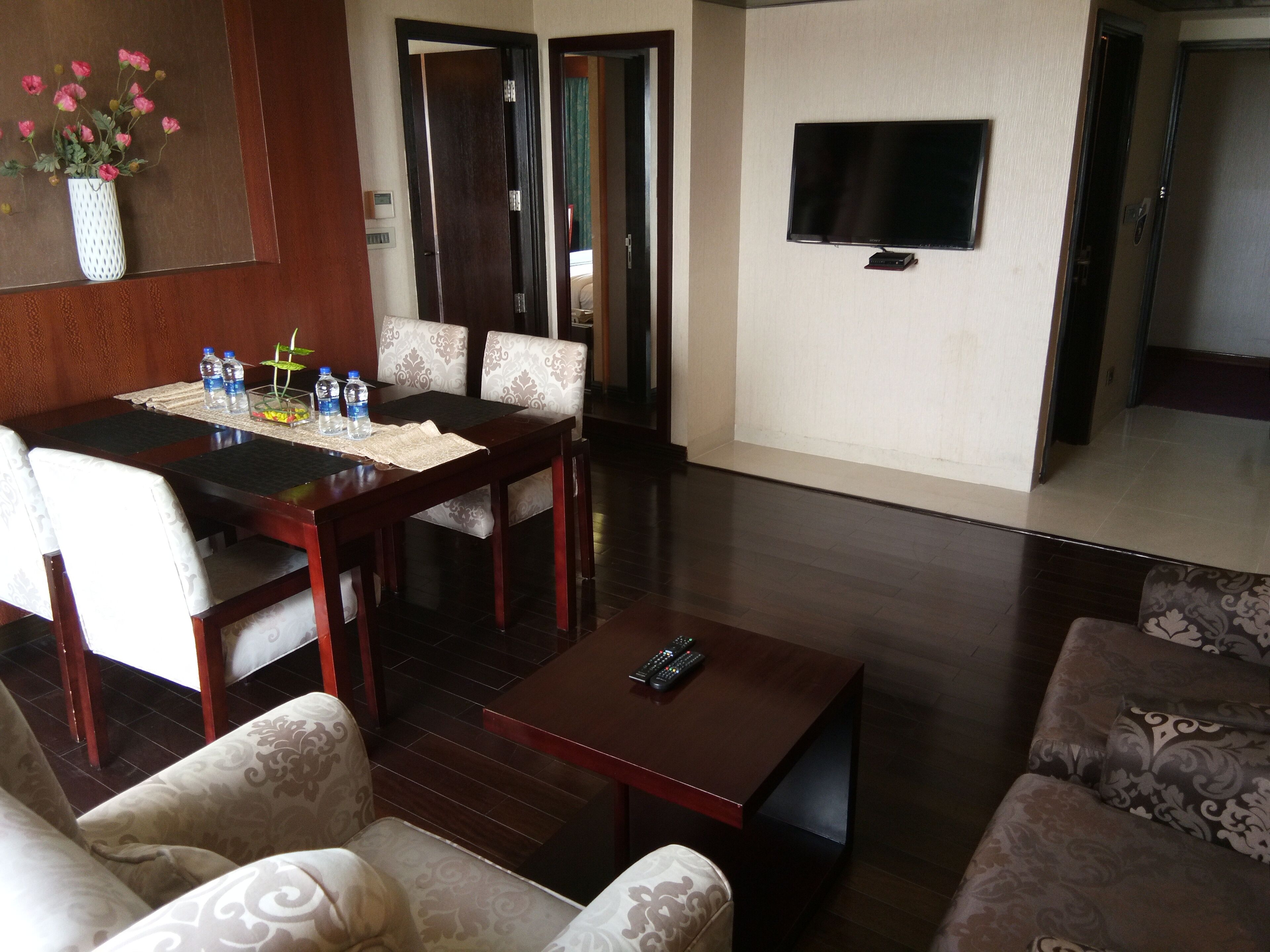 A living room with a dining table, chairs, a sofa, a coffee table, a TV, and a vase with flowers.