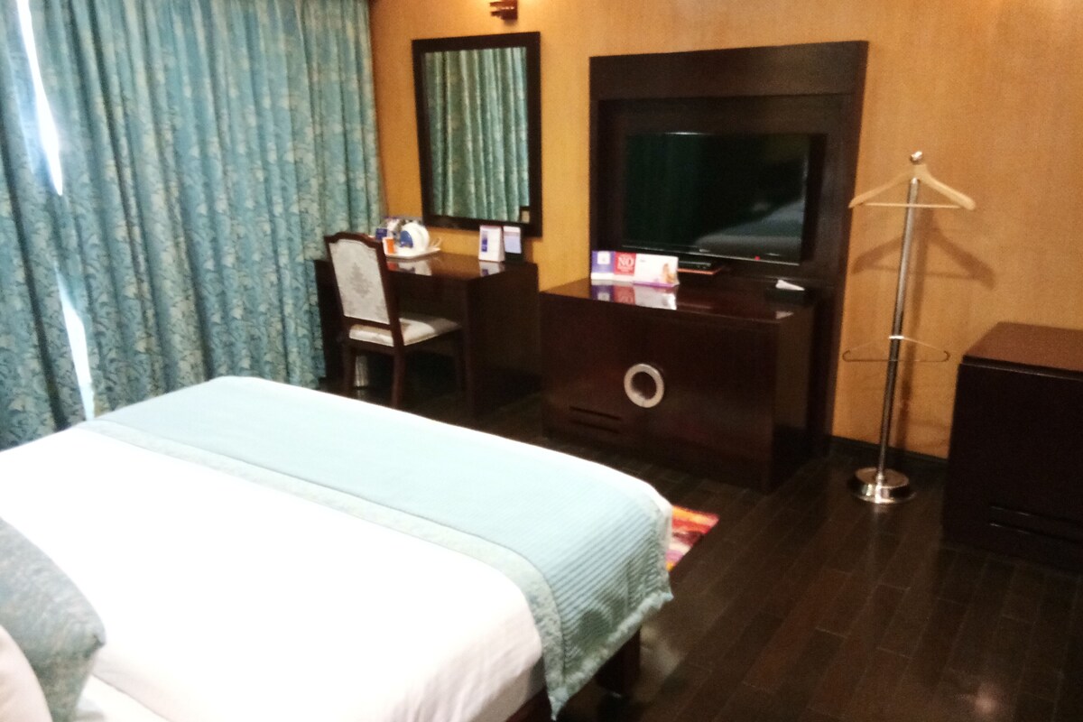Habitación de hotel con cama, escritorio, silla, televisor y lámpara de pie.
