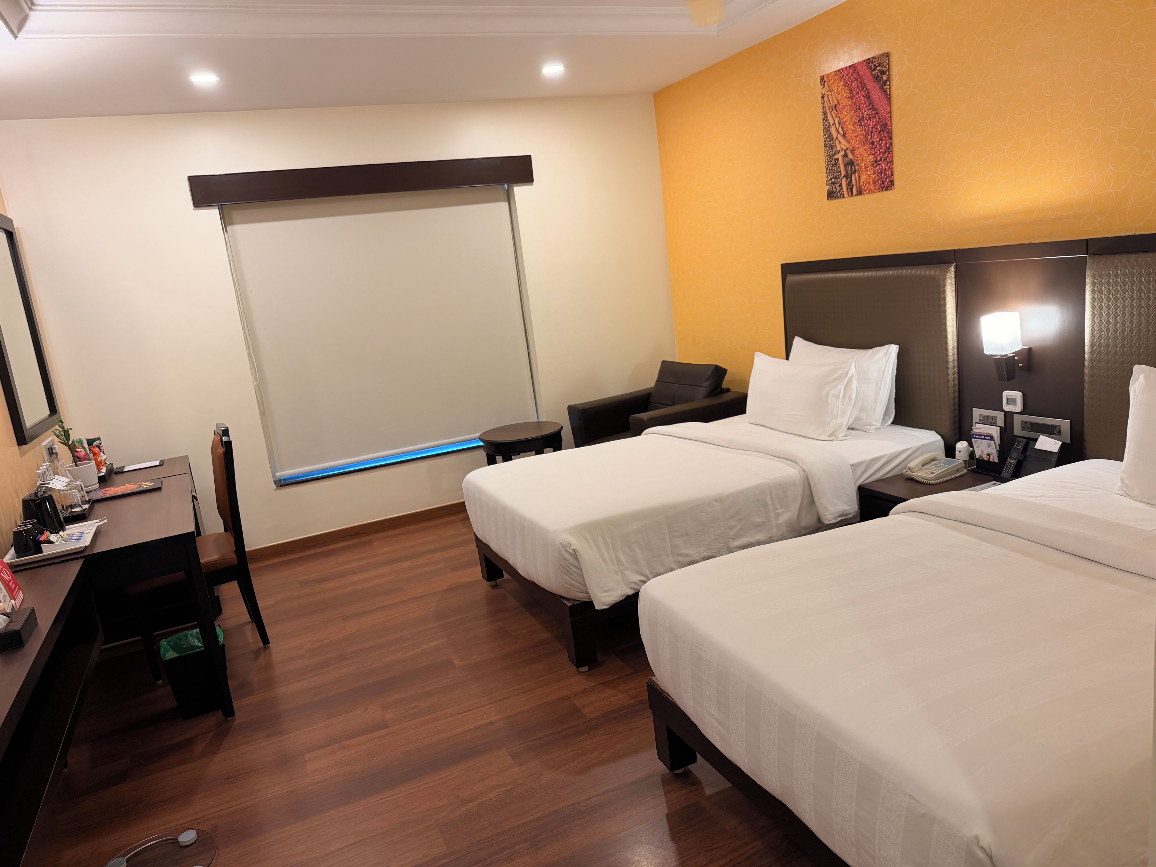 A hotel room with two beds, a desk, a chair, a small table, a bedside table with a phone, and a wall-mounted lamp.