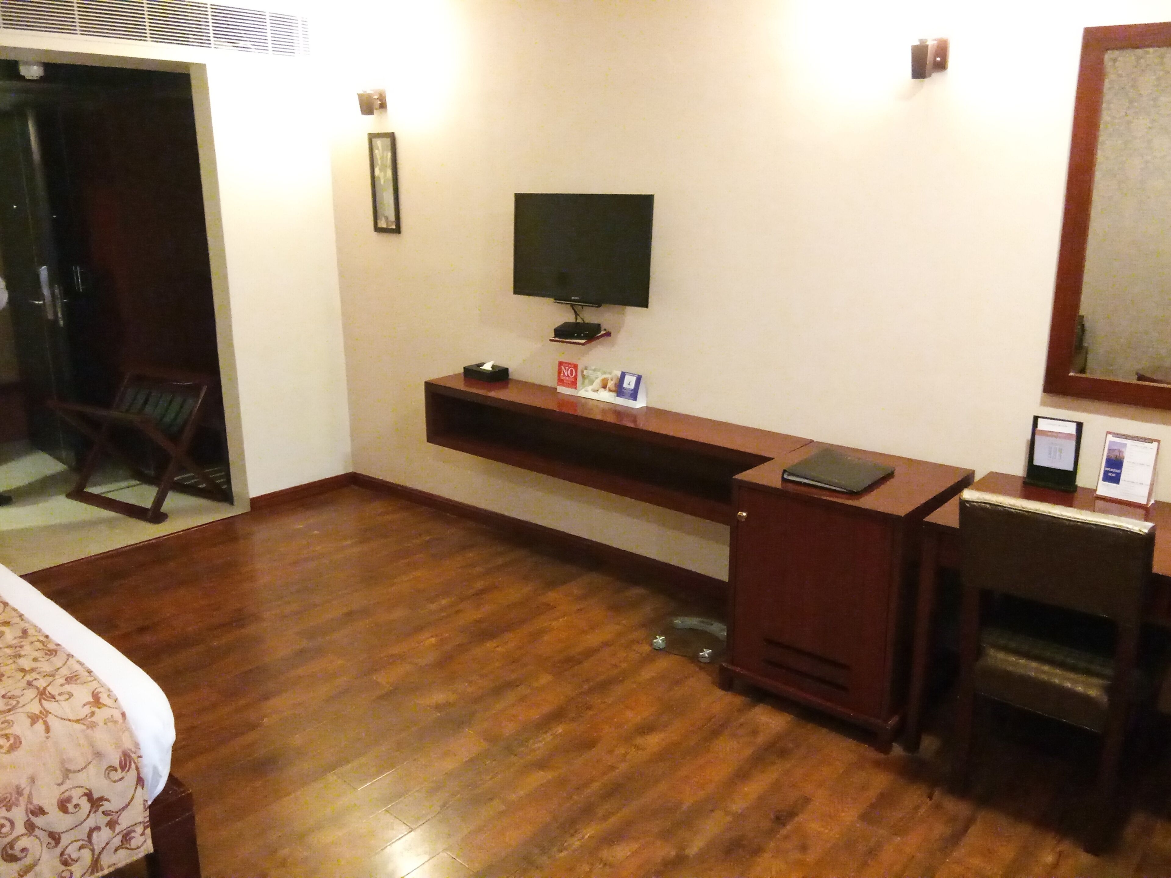 A hotel room with a wooden floor, a bed with a patterned bedspread, a desk with a computer, a television, and a chair.