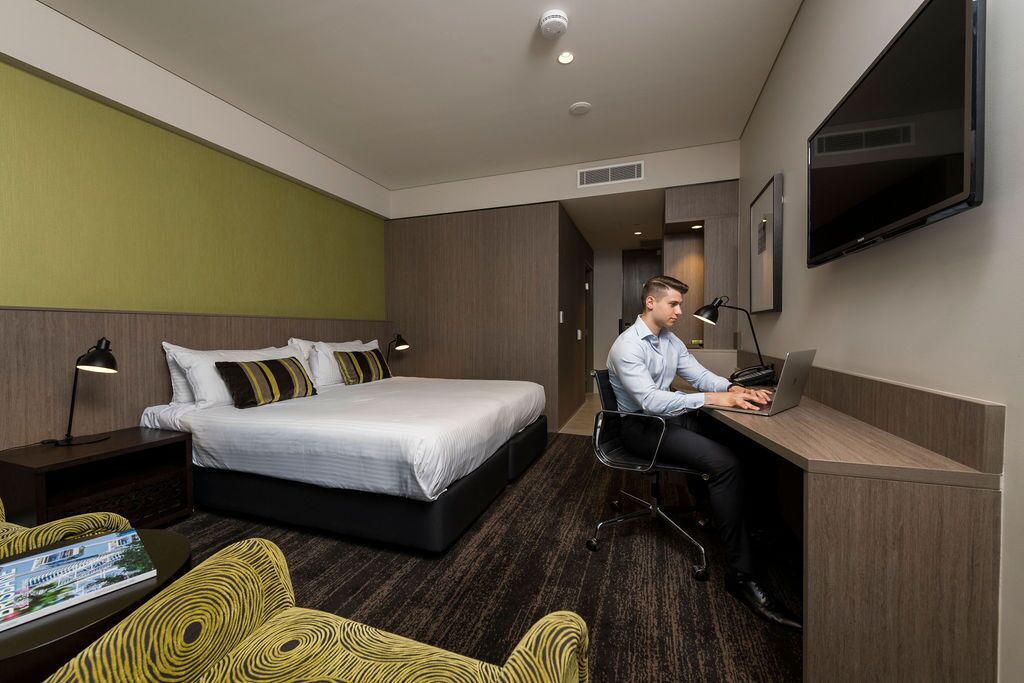 Habitación de hotel con cama, escritorio con una computadora portátil, silla, televisor y mesita de noche con lámpara.