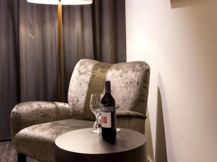 Habitación de hotel con un sillón cómodo, una mesita redonda con una botella de vino y una copa, una lámpara de pie y un cuadro en la pared.