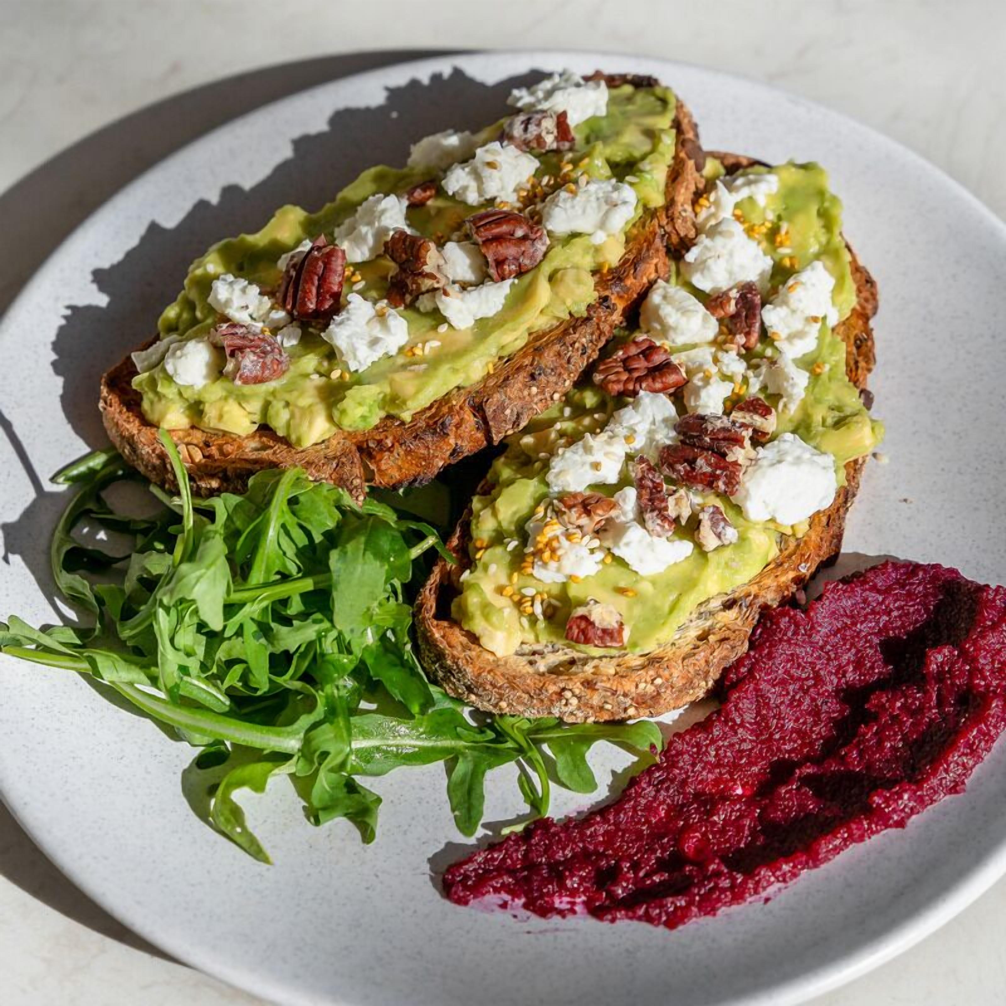 A plate with avocado toast topped with pecans and feta cheese, accompanied by a side of arugula and beetroot puree.