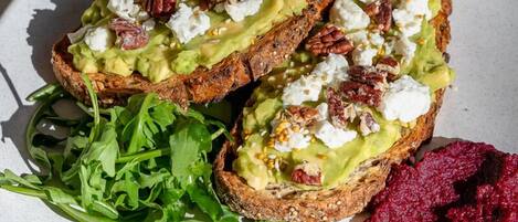 Un plato con tostada de aguacate, nueces y queso feta, acompañado de rúcula y puré de remolacha.