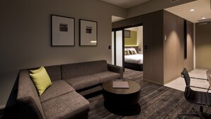 Una habitació d'hotel moderna amb un sofà, una taula de centre rodona i una cadira. Es veu un llit a través d'una porta oberta, i la habitació té una paleta de colors neutres amb quadres a la paret.