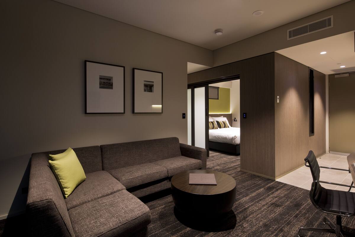 A modern hotel room with a sofa, a round coffee table, and a single chair. There is a bed visible through an open door, and the room has a neutral color scheme with framed pictures on the wall.