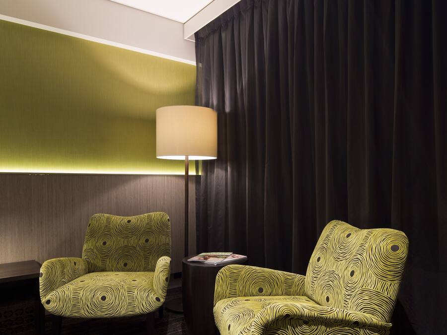 A hotel room with two green patterned armchairs, a side table with a lamp, and a dark curtain backdrop.