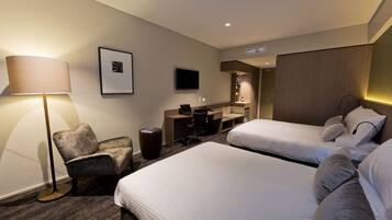 A hotel room with two beds, a desk, a chair, a TV, and a lamp.