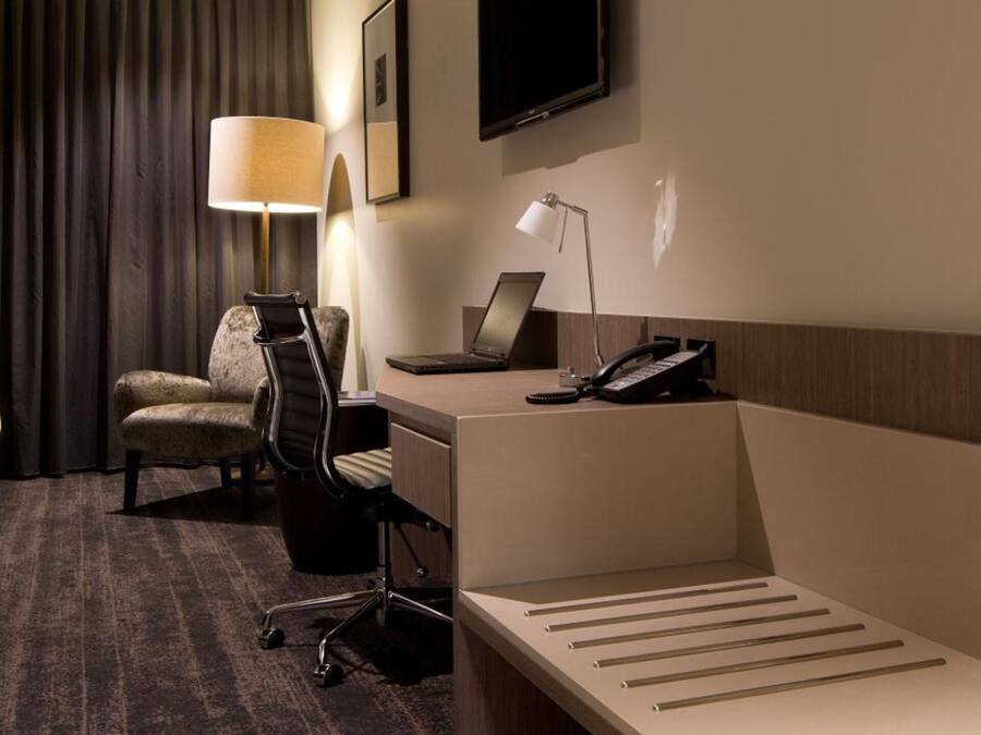 A hotel room with a desk, chair, lamp, and a flat-screen TV mounted on the wall.