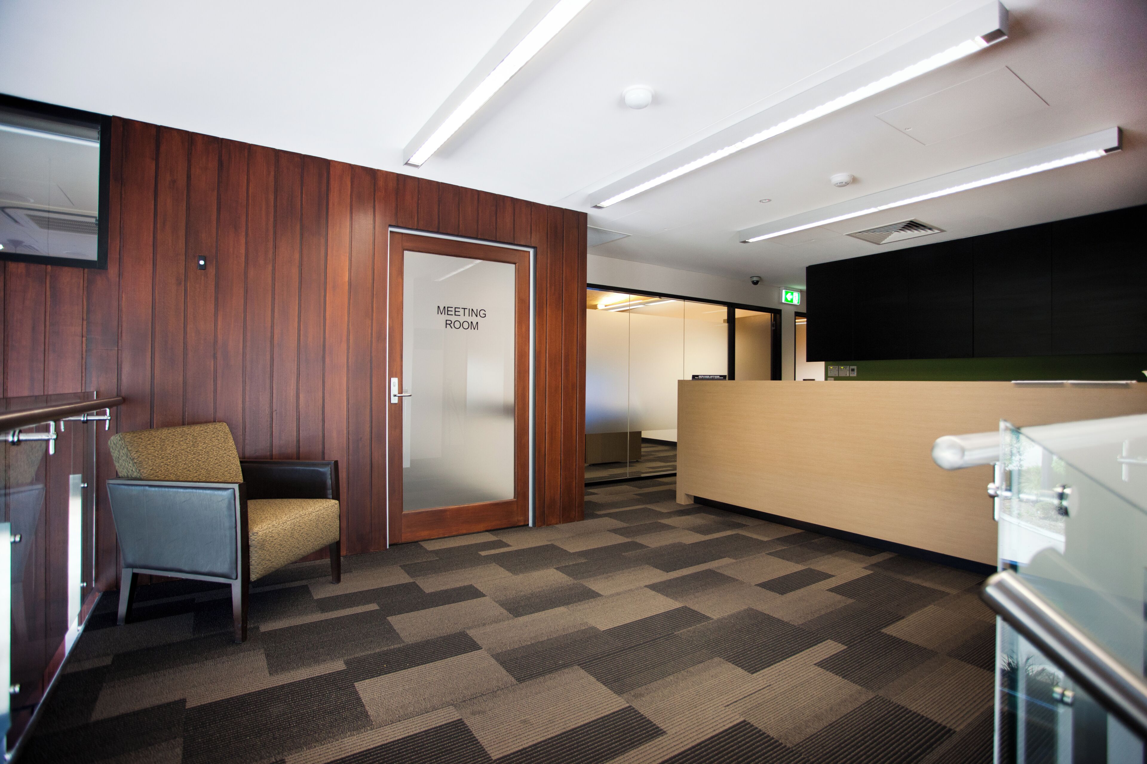 A modern meeting room with a wooden panel wall, a glass door labeled 'MEETING ROOM', a reception desk, and a patterned carpet.