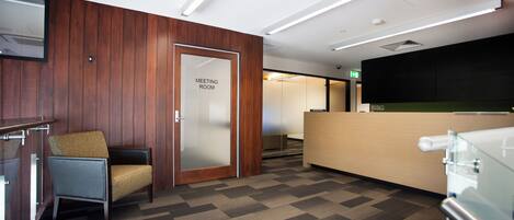 A modern meeting room with a wooden panel wall, a glass door labeled 'MEETING ROOM', a reception desk, and a patterned carpet.