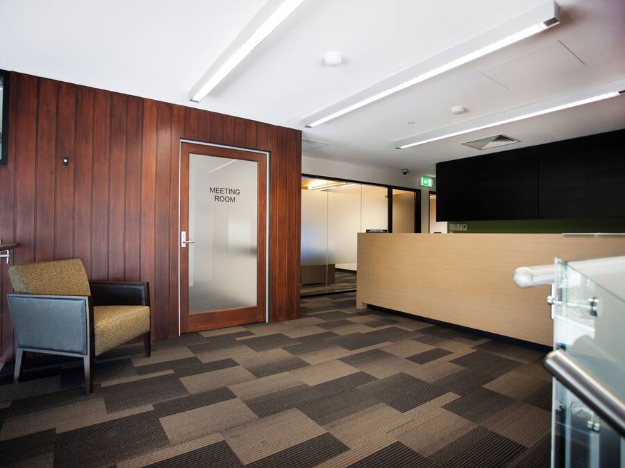 A modern meeting room with a wooden panel wall, a glass door labeled 'MEETING ROOM', a reception desk, and a patterned carpet.