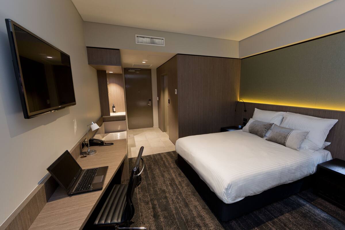 A modern hotel room with a large bed, a desk with a laptop, a TV mounted on the wall, and a separate bathroom.