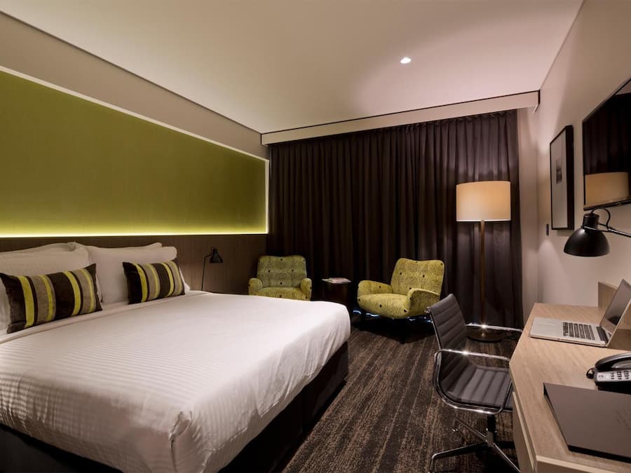 A hotel room with a large bed, two armchairs, a desk with a laptop, and a desk lamp.