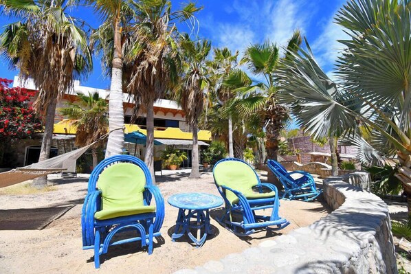 A beachside lounge with blue and green lounge chairs and a small round table, surrounded by palm trees and a building with a yellow awning.