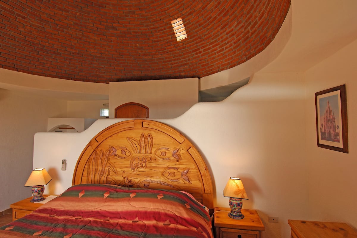 A bedroom with a wooden headboard, two bedside lamps, a bed with a colorful blanket, a brick ceiling, and a framed picture on the wall.