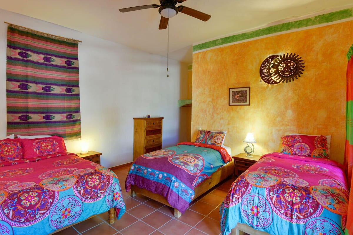 A room with two beds, a ceiling fan, a wall with a sun decoration, and a colorful tapestry.
