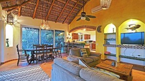 A spacious living area with a thatched ceiling, a dining set, a sofa, a TV, and a view of the outdoors. - Ventana Bay Resort (El Sargento)
